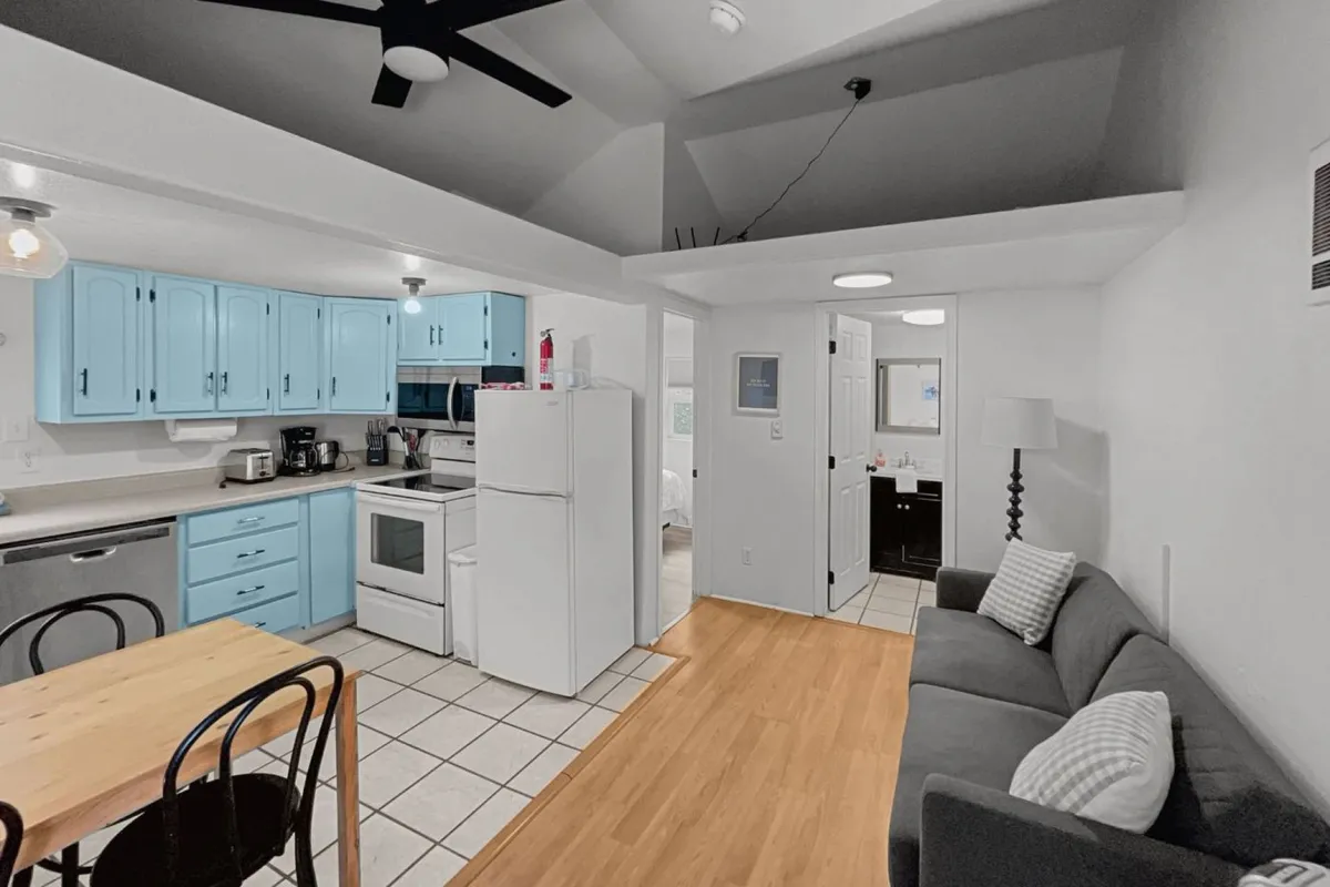 This image captures the main living area, featuring blue kitchen cabinets, a dining table, and a sofa in a single open space. The scene clearly shows how the kitchen, dining, and living functions integrate, with ample natural light and a cozy, inviting setup for relaxing or dining together.