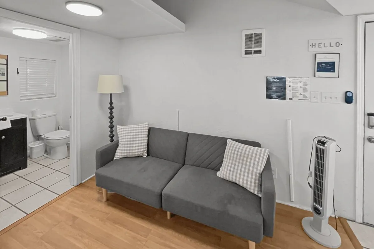 A cozy living room with a gray couch, floor lamp, wall décor, and a door leading to another area, all bathed in natural light. The mood is casual and welcoming, ideal for relaxing after coastal adventures or enjoying intimate conversations.
