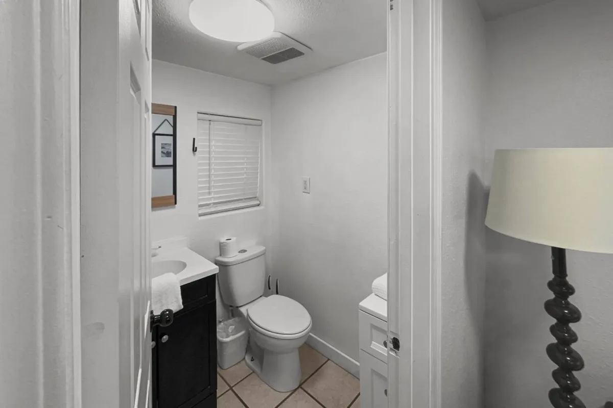 A minimalistic, bright bathroom with a modern sink, toilet, lamp, and natural light filtering through a window. The space feels clean and uncluttered, ideal for refreshing and preparing for the day.