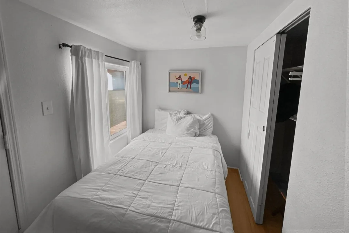 A serene bedroom scene featuring a full bed with crisp white bedding, framed artwork on the wall, and a window with sunlight streaming through sheer curtains. The atmosphere is calm and welcoming, ideal for restful nights and leisurely mornings.