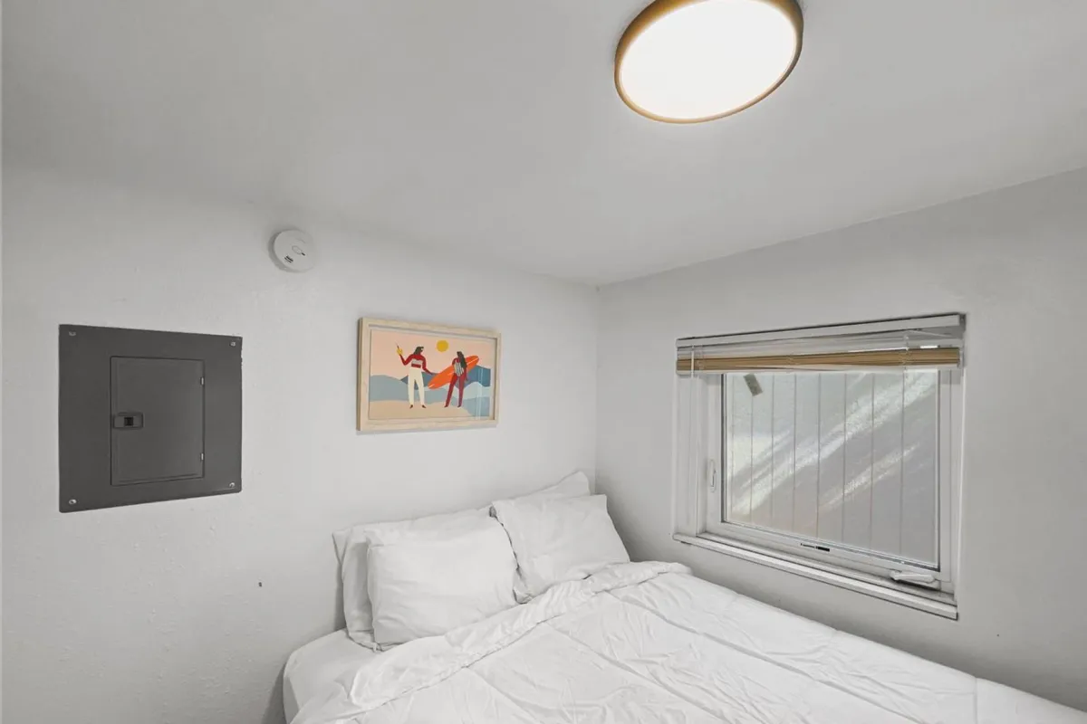 The image showcases a cozy bedroom interior with a comfortable bed bathed in natural light from a large window, inviting artwork on the wall, and a peaceful, uncluttered atmosphere ideal for relaxation.