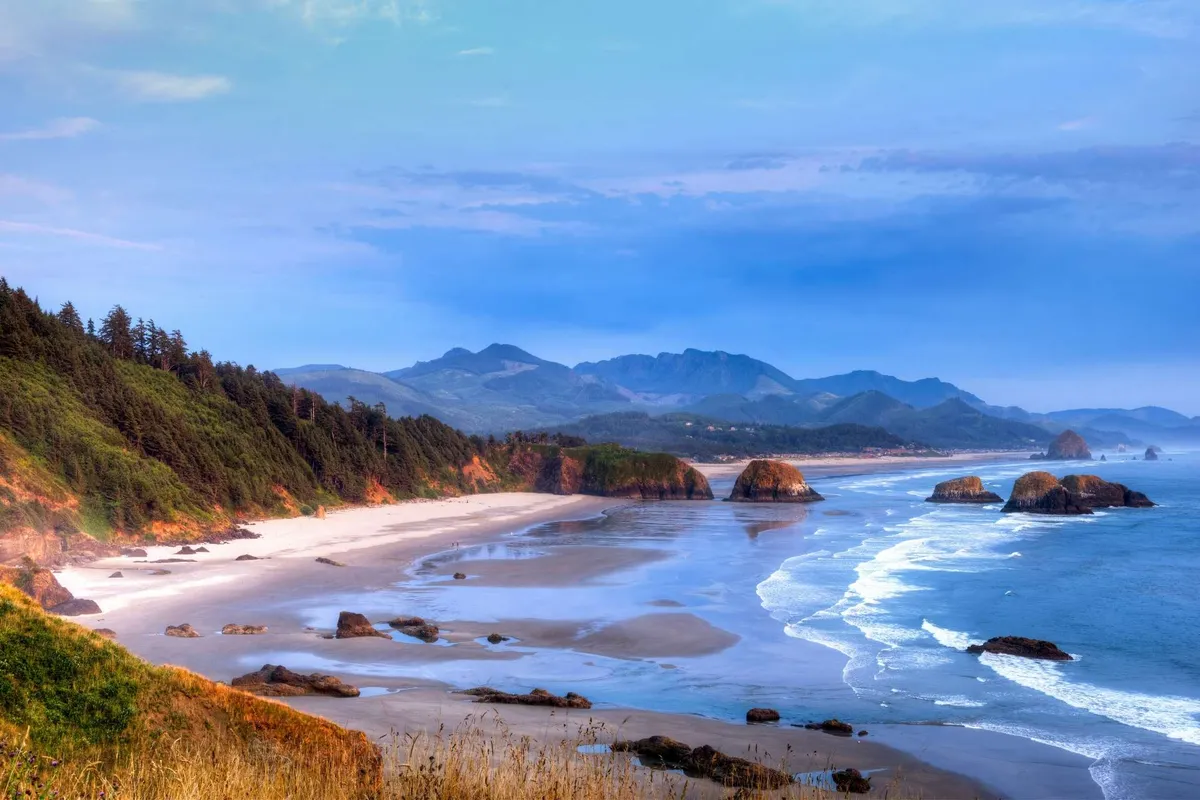 A tranquil panoramic view of the beach with ocean waves crashing against dramatic rock formations, framed by a mountain backdrop. The foreground offers ample space for gatherings and relaxation, evoking the communal spirit of outdoor living.
